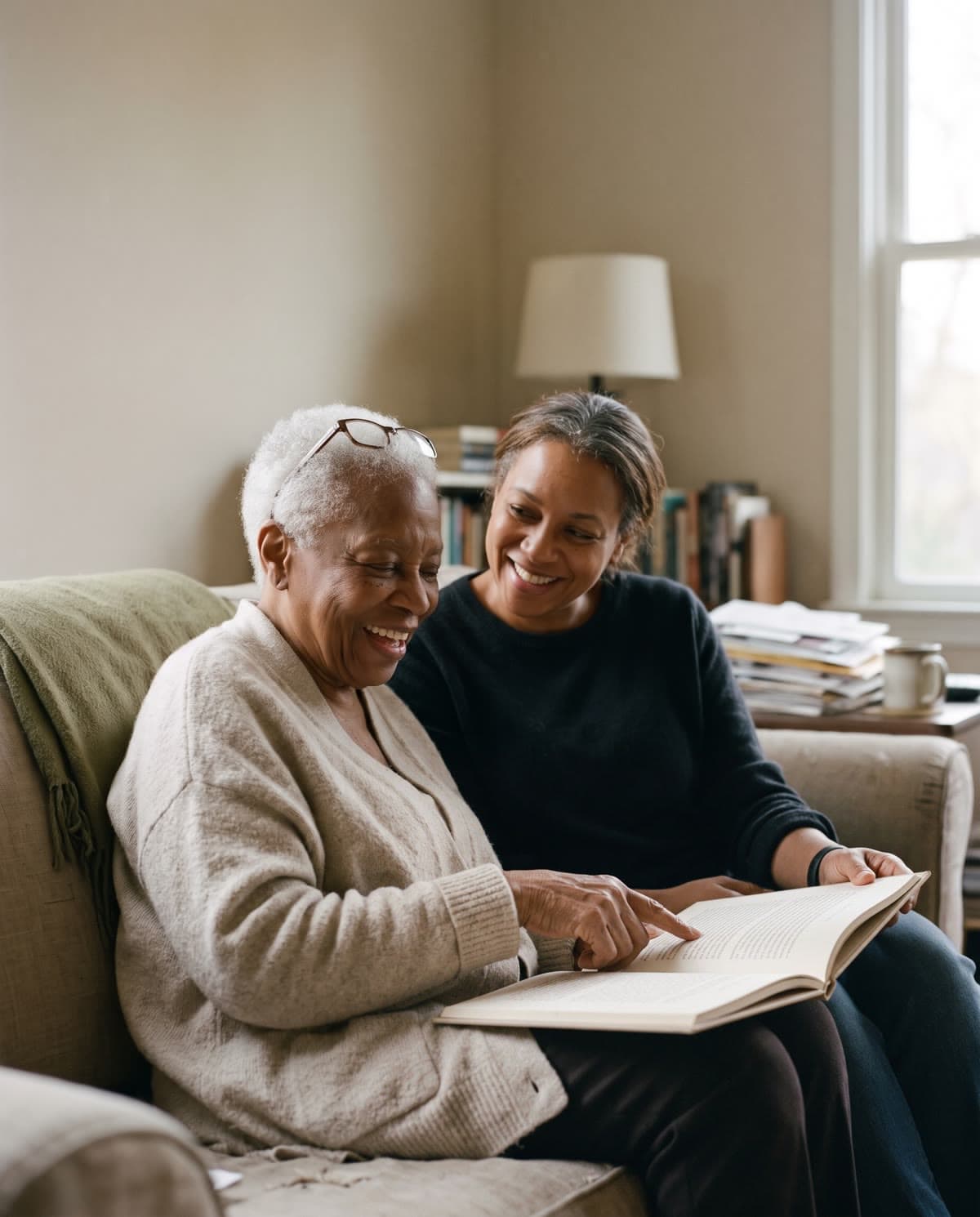 An older woman laughs as she and her grown daughter read a book together on the sofa. AI-generated image.