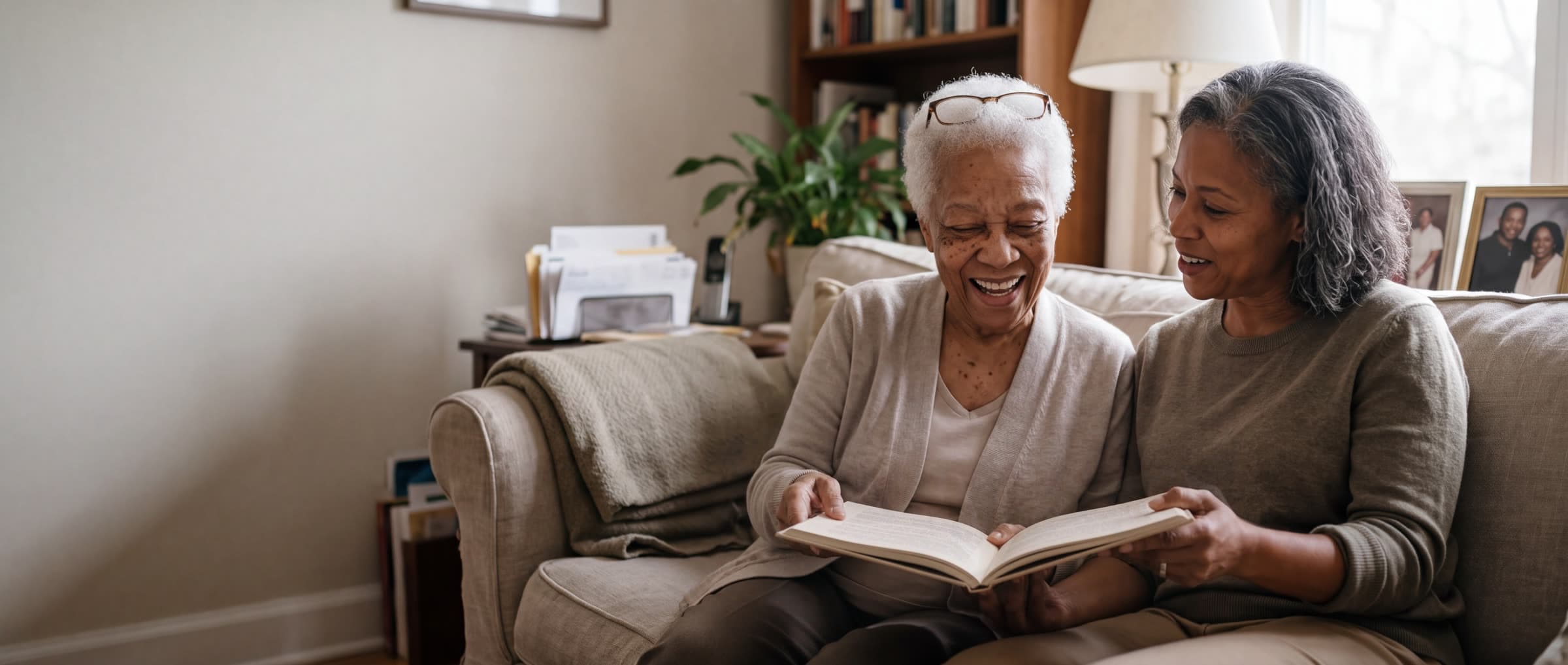 An older woman laughs as she and her grown daughter read a book together on the sofa. AI-generated image.