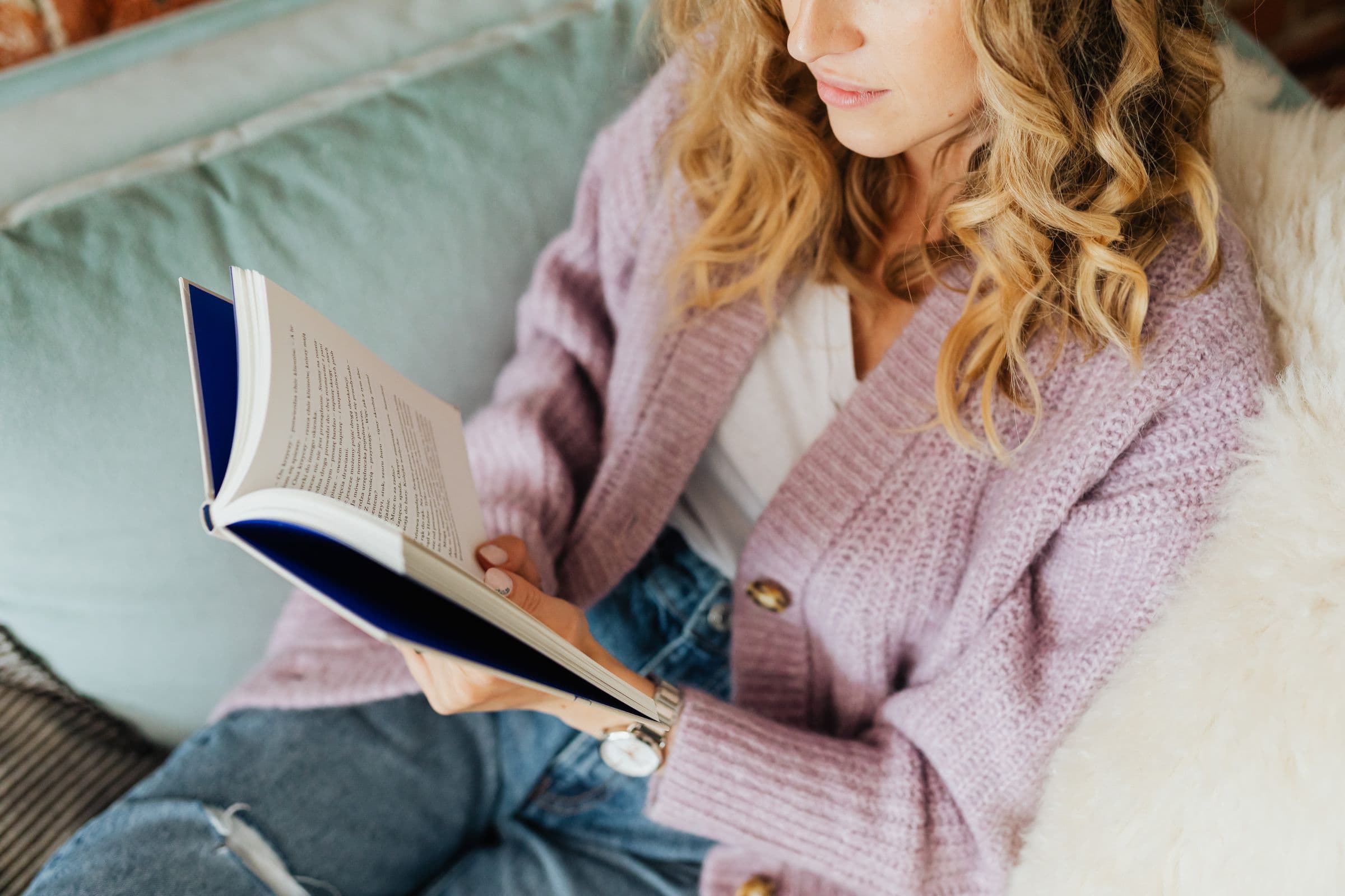 A woman reading a book on a sofa, in a lilac cardigan.