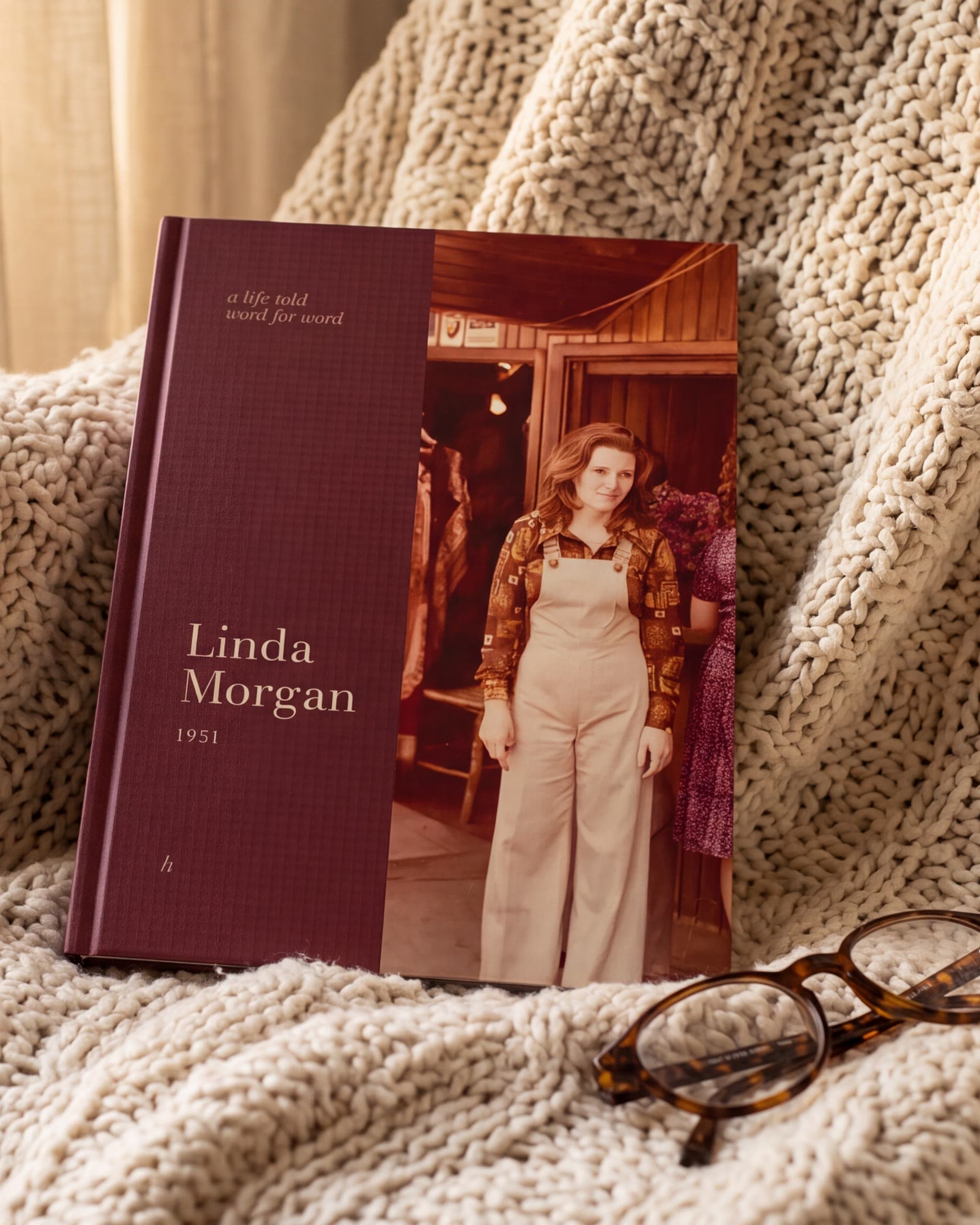 A slim hardcover memoir with an old colour photo of a young woman in overalls on its burgundy cover, resting on a knitted blanket on an armchair, next to a pair of reading glasses. AI-generated image.